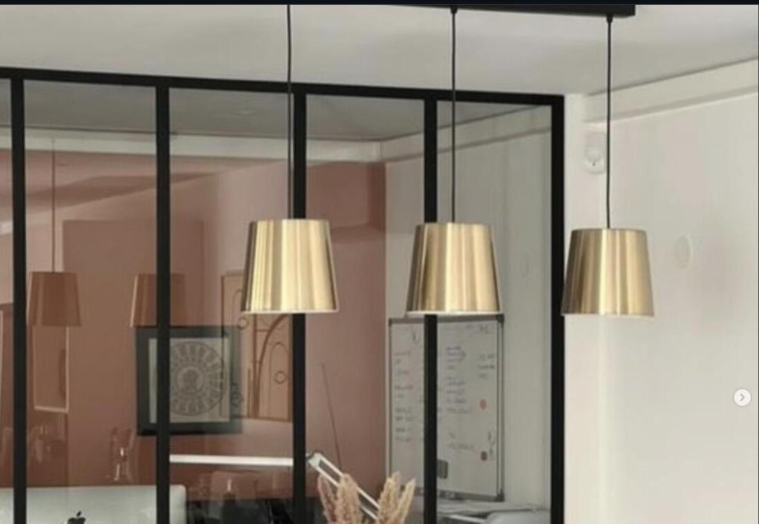 Verrière en acier noir séparant un bureau et une salle à manger, suspensions lumineuses dorées au premier plan.