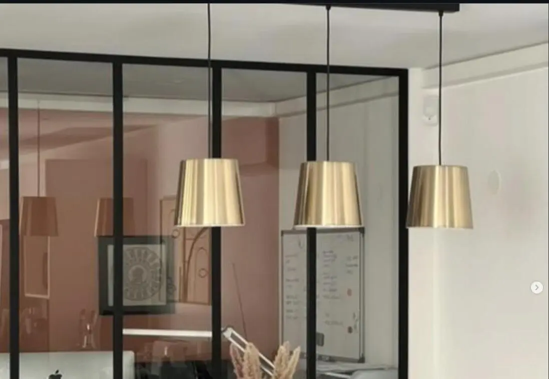 Verrière en acier noir séparant un bureau et une salle à manger, suspensions lumineuses dorées au premier plan.