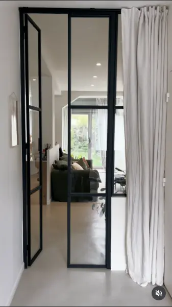 Verrière à porte pivotante et profilés noirs fins, ouverte sur un salon lumineux avec rideau en lin.