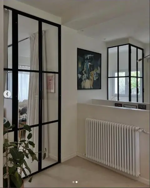 Verrière métallique noire à double porte vitrée, ouverte sur un salon avec tableau mural et radiateur design.